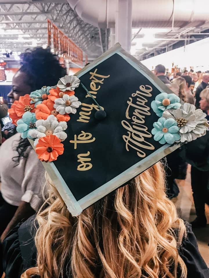 back of woman's head at a graduation showing graduation cap that has blue and coral flowers with the words "on to my next adventure" in the center