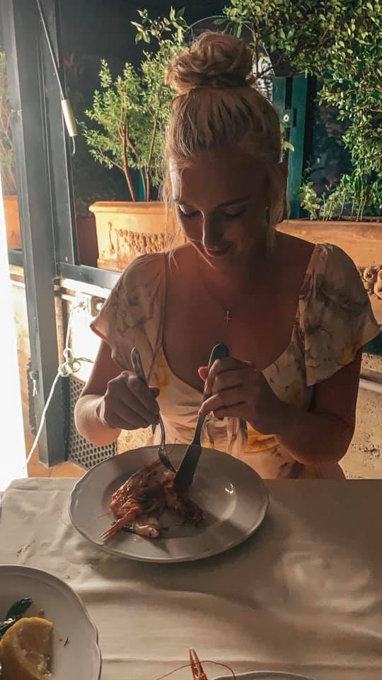 A woman in a pink floral dress at a restaurant for a late dinner attempting to cut the seafood on her plate.