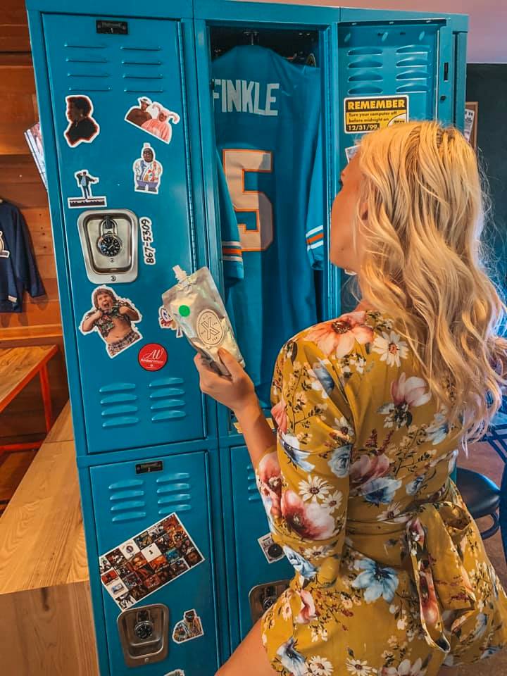 A woman in a yellow floral dress holding an adult capri sun looking into a bright blue locker