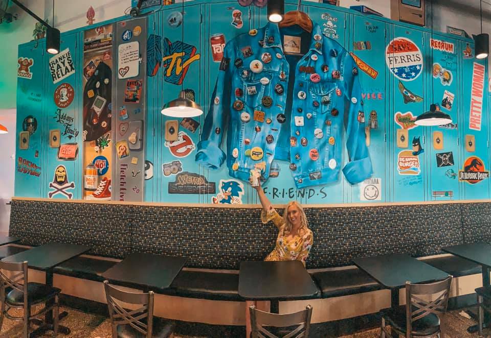 Woman holding up an adult Capri Sun in front of an '80 and '90s themed locker mural.  Includes references to MTV, Friends, Ferris Buehler's Day Off, and others.