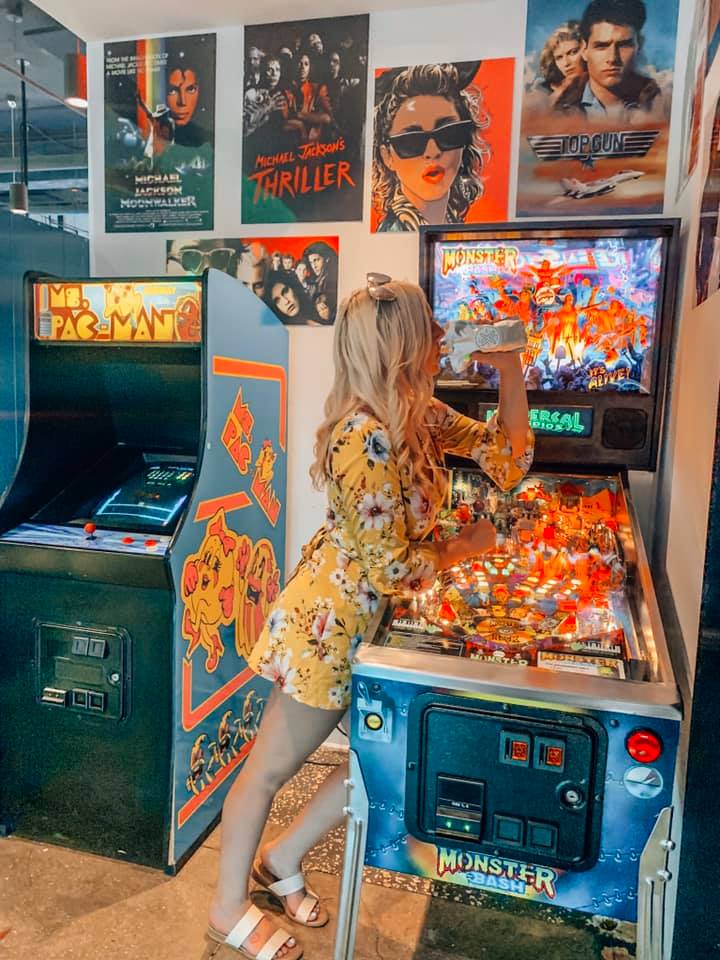 a woman in a yellow floral dress leaning against a pinball machine drinking an adult capri sun