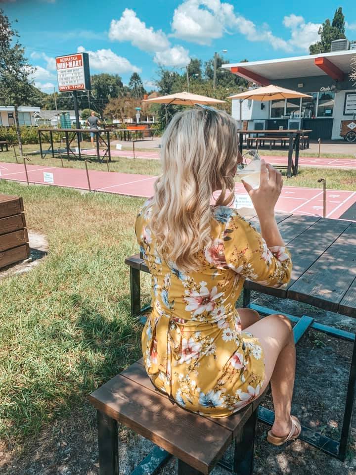 a woman in a yellow dress sipping on a glass of white wine at a picnic table at Nebraska Mini Mart