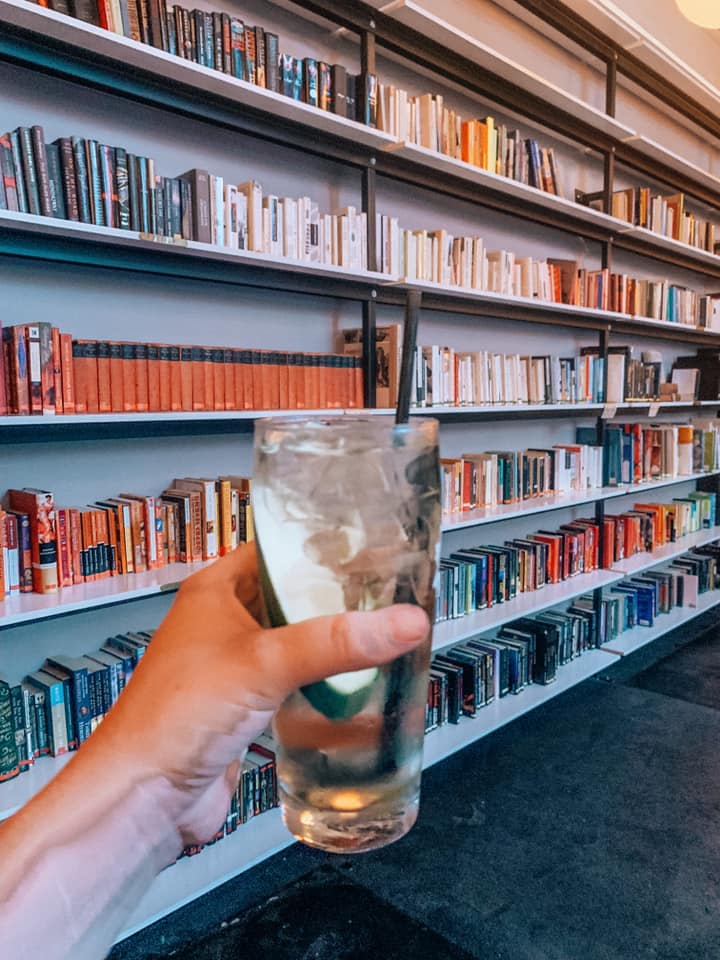 a woman holding a drink in front of a colorful rainbow coordinated bookshelf