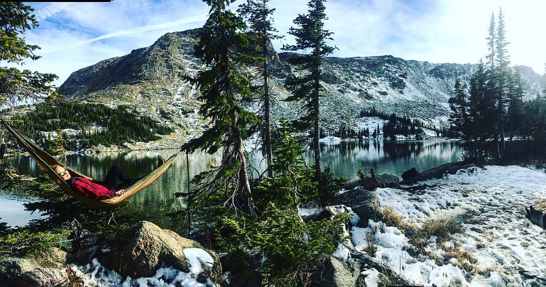 Woman laying hammock on the top of Colorado mountains surrounded by trees and a lake. This is how to travel more!