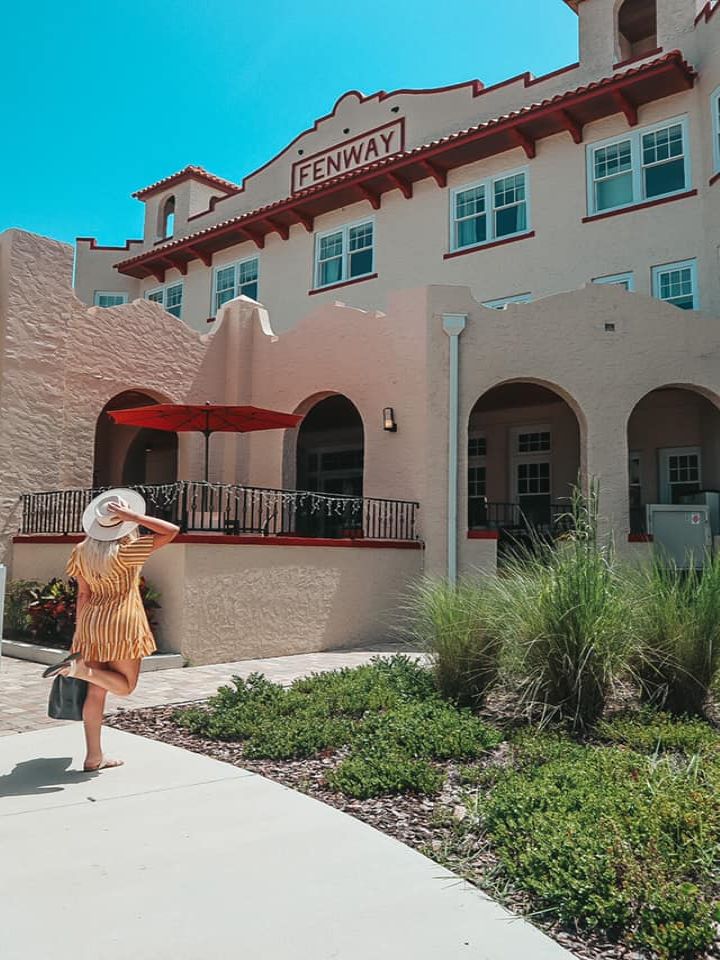 woman kicking her leg up and holding her hat and suitcase while looking at the beautiful Fenway Hotel in Dunedin, Florida