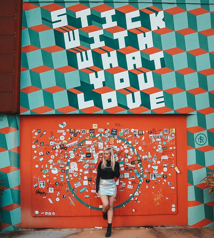 Woman leaning up against a mural in downtown St. Pete that says "Stick with what you love"