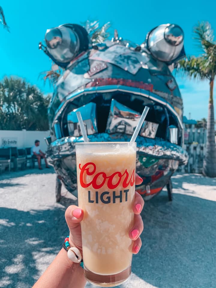 Hand holding a pina colada up in front of a giant metal fish at The Ugly Grouper