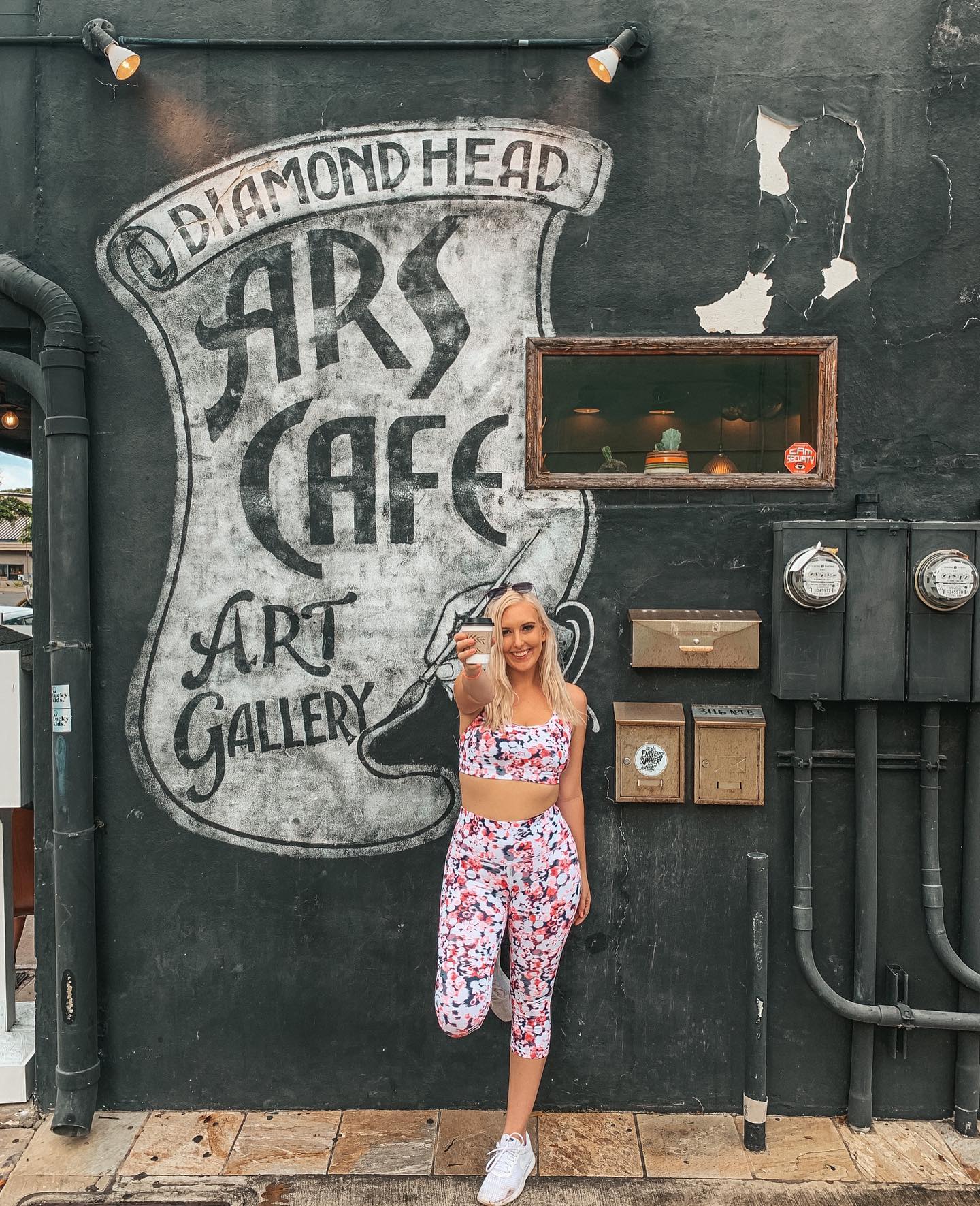 Woman wearing a pink speckled workout outfit and holding up coffee outside of Ars Cafe in front of their mural that reads "Diamond Head Ars Cafe Art Gallery"