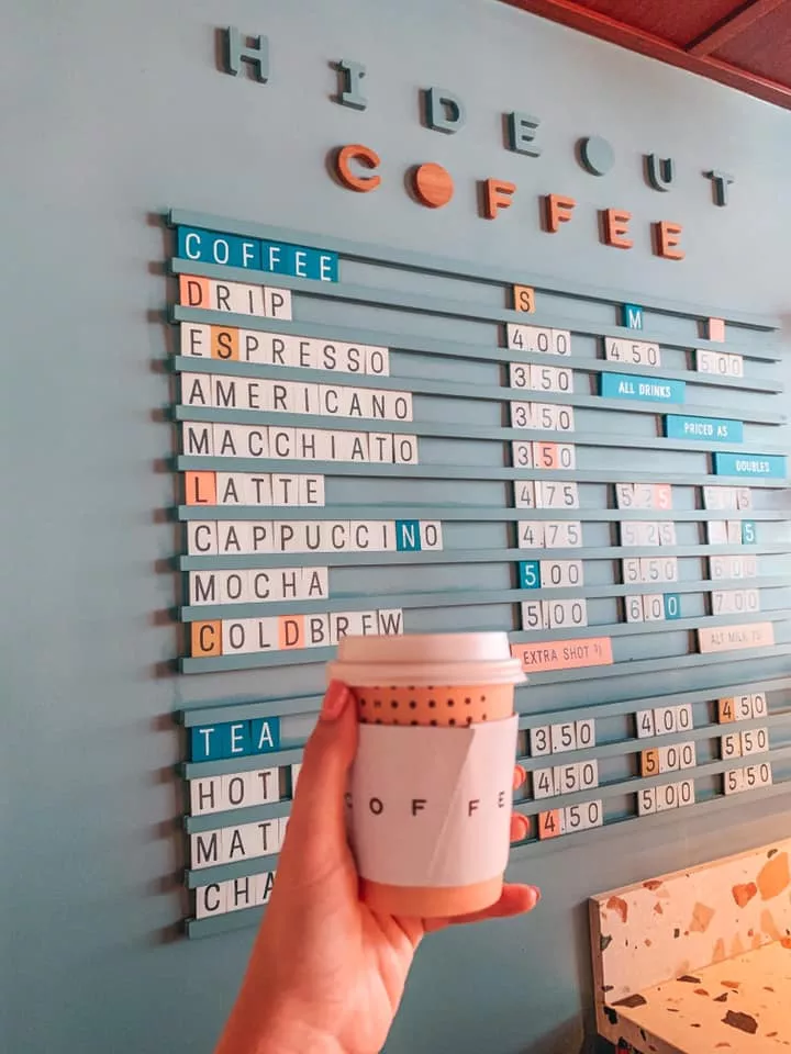 Holding up coffee cup in front of the menu at Hideout Coffee