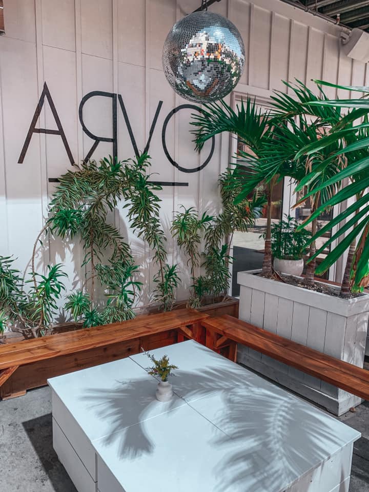 Outdoor seating area at Arvo. Brown benches, a beautiful white table, plenty of hanging and sitting plants, as well as a disco ball.