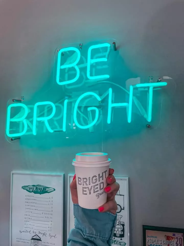 Holding up coffee from bright eyed brew co in front of a neon sign that reads "be bright"