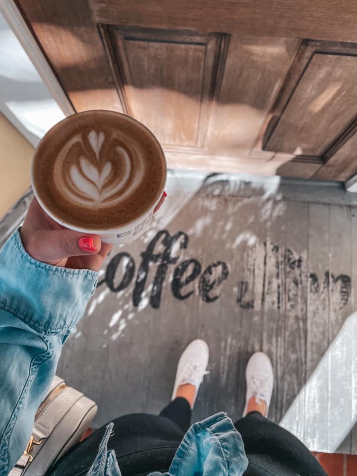 holding latte with fun design over doorstep area that reads 