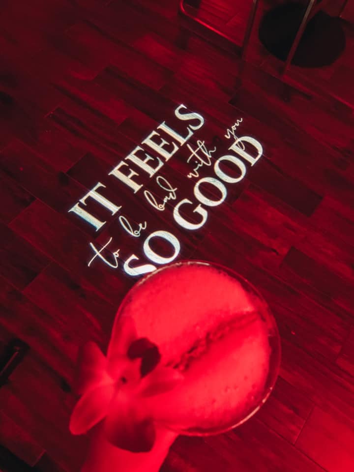 holding "sloth" drink over a quote being projected on the floor with red lighting "It feels so good to be bad with you"