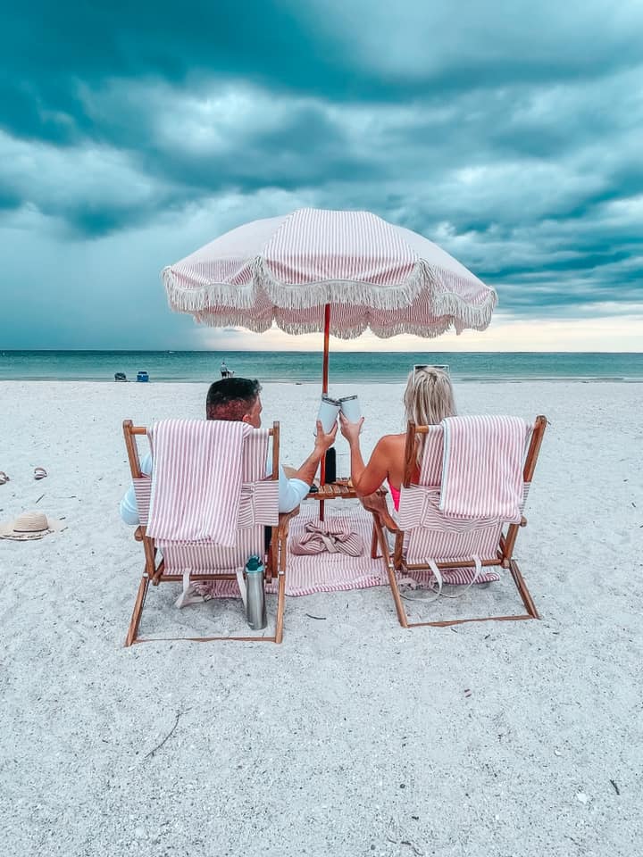 sitting under pink and white striped umbrella cheersing mugs for a beach day date