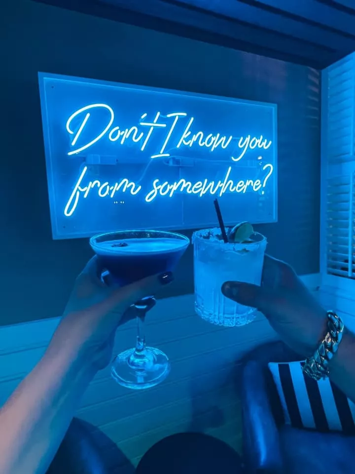 martini and margarita cheersing in front of a blue neon sign that says 