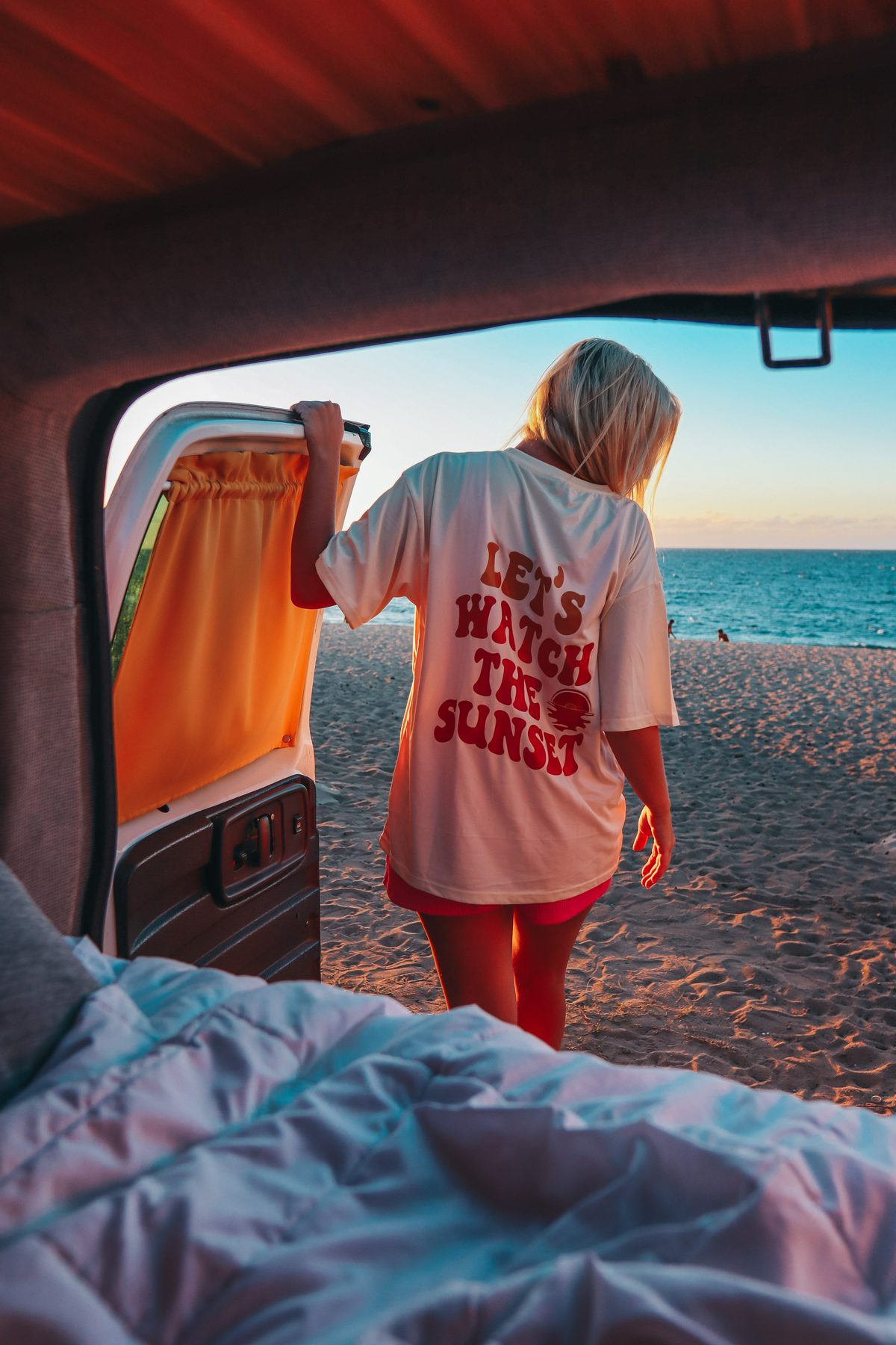 Woman wearing a "let's watch the sunset" tee hanging outside of a van doing van life on Oahu