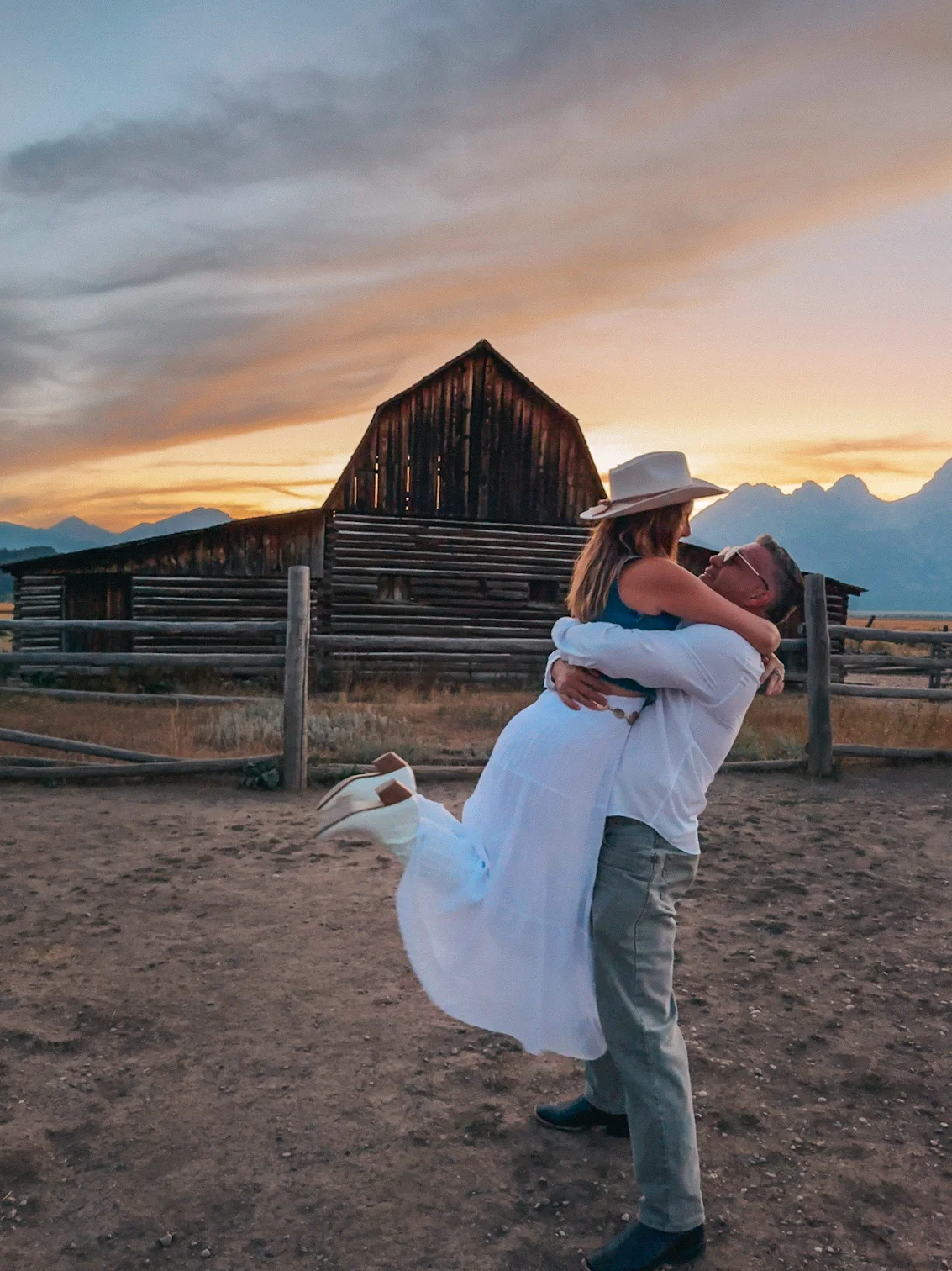 Man Lifting Up A Women With Her Feet Kicked Up Behind Her In Front Of Mormon Row In Wyoming