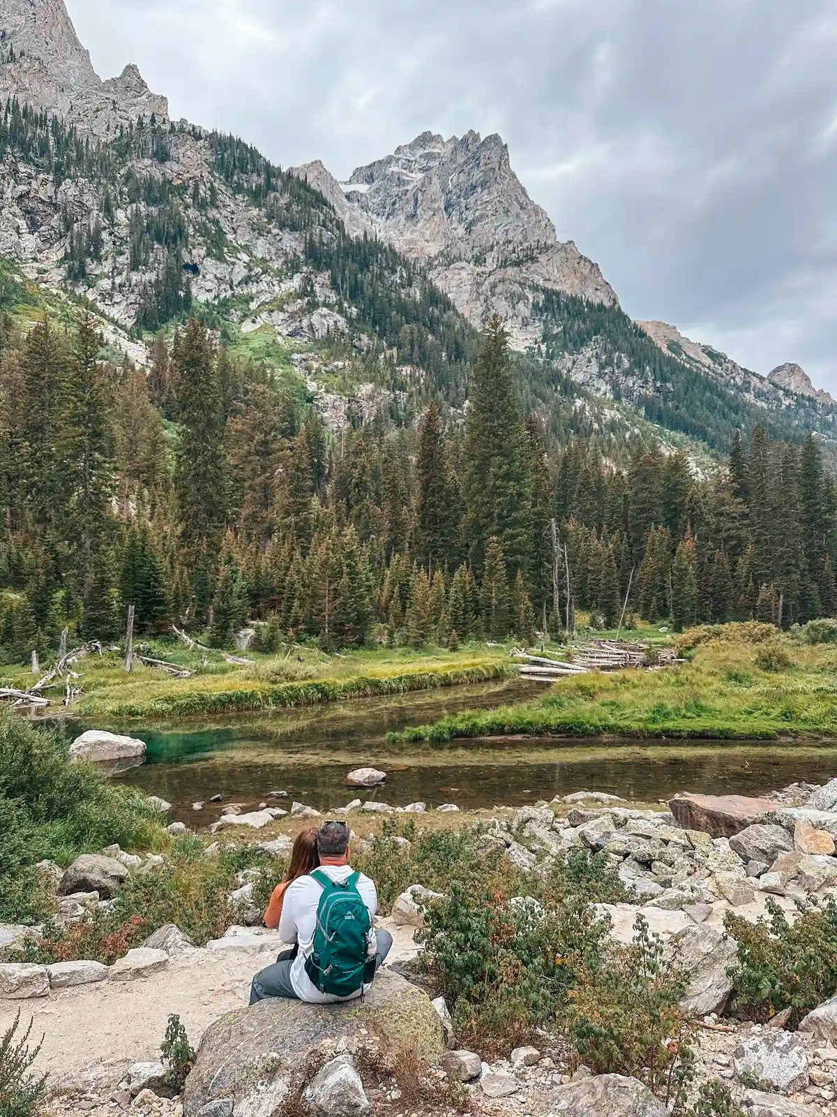 Couple Sitting On A Rock Looking Out At Stream And Mountains On The Cascade Canyon Trail In Grand Teton National Park